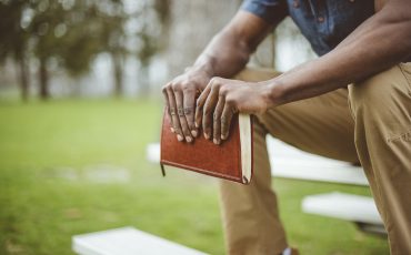 closeup-shot-male-holding-bible-while-sitting-park-table